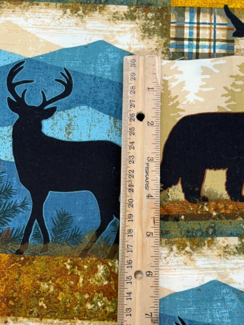 May include: Fabric with a nature-themed design featuring a black silhouette of a deer with antlers, a bear, and mountain scenery. The fabric is measured with a wooden ruler, showing a length of approximately 6 inches.