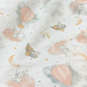 May include: A white fabric with a whimsical pattern featuring bunnies, hot air balloons, crescent moons, clouds, and stars in soft pastel colors. The design includes peach, gray, and gold accents, creating a dreamy, celestial theme.