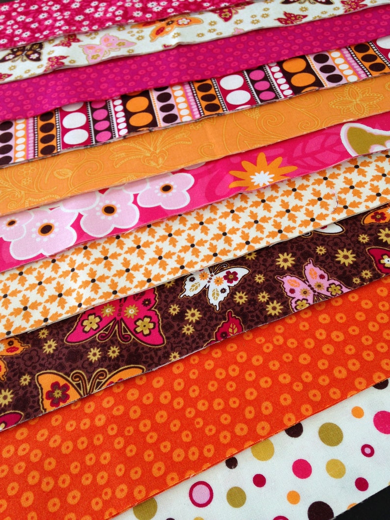 May include: A stack of colorful fabric scraps with various patterns, including polka dots, butterflies, and floral designs. The fabrics are in shades of pink, orange, brown, and white.
