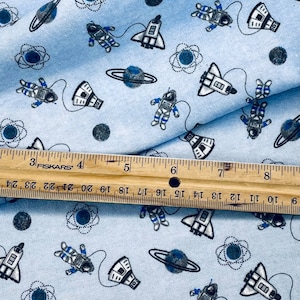 May include: Blue cotton fabric with a repeating pattern of white astronauts, spaceships, planets, and atoms. The fabric is folded in half and a ruler is placed on top of the fabric.