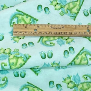 May include: Light blue fabric with a repeating pattern of green dragons and blue footprints. The fabric is folded over and a wooden ruler is placed on top of the fabric.