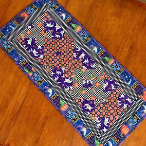May include: A rectangular Halloween table runner. The runner has a purple border with Halloween icons, a black and white checkered pattern, and a central design with purple ghosts and orange pumpkins. The runner is made of fabric.