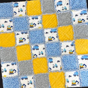 May include: A patchwork baby blanket featuring squares of blue, yellow, white, and gray fabrics. The design includes construction vehicles like dump trucks and excavators. The blanket has a soft, textured appearance.