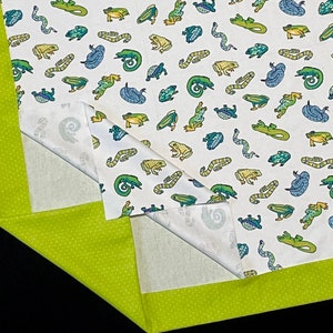 May include: A white fabric with a green border and a repeating pattern of green, blue, and brown frogs and lizards. The fabric is folded in half, showing the pattern on both sides.