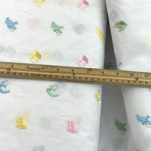 May include: White fabric with a repeating pattern of colourful embroidered blocks and animals. The fabric is measured with a ruler.