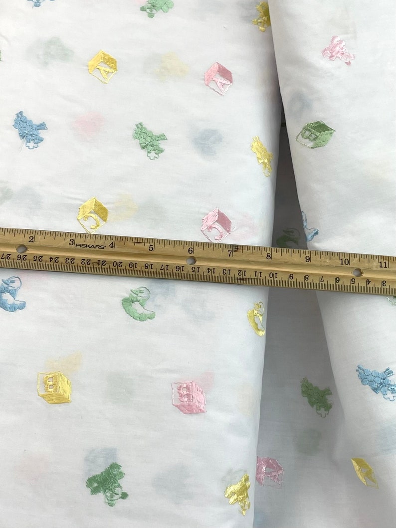 May include: White fabric with a repeating pattern of colourful embroidered blocks and animals. The fabric is measured with a ruler.