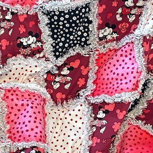 以下が含まれることがあります： ミッキーマウスとミニーマウスをテーマにしたピンクと赤のパッチワークキルト。キルトは、水玉模様、ハート、花など、さまざまなパターンを持つさまざまな生地で作られています。キルトは白いレースで縁取られています。