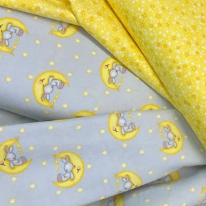 May include: Two pieces of fabric with a repeating pattern of bunnies sleeping on a crescent moon. One fabric is light blue with yellow stars and the other is yellow with white stars.
