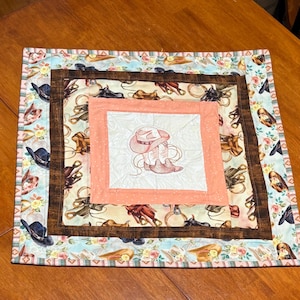 Peut inclure: Un set de table matelassé sur le thème du cow-boy. Le set de table présente un carré central avec un motif brodé d'un chapeau et de bottes de cow-boy. Le motif est entouré d'une bordure couleur pêche, puis d'un carré avec un tissu sur le thème du cow-boy.
