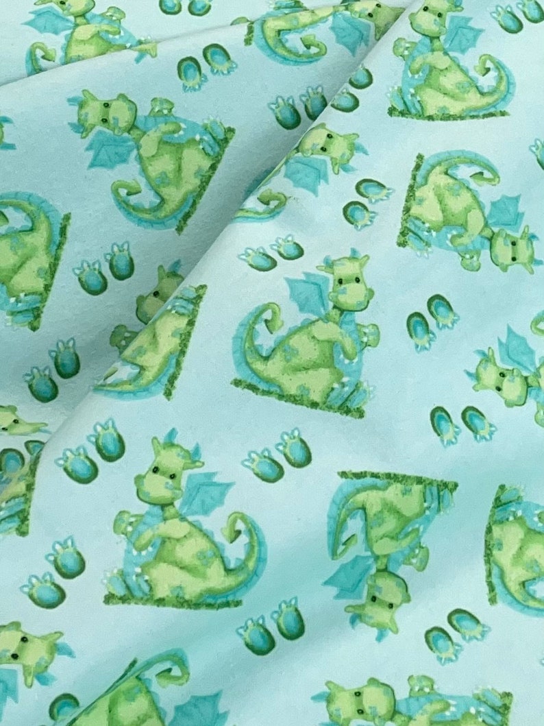 May include: A light blue fabric with a repeating pattern of green dragons and blue footprints. The dragons are cartoon-like and have wings and tails.