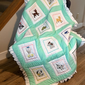May include: A handmade baby quilt with a mint green and white color scheme. The quilt features embroidered animal designs within square blocks, including a goat, horse, and dog. The quilt is trimmed with a white ruffled edge.