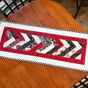 May include: A rectangular table runner with a festive design. The runner features a red center with white and black chevron patterns, accented by a black and white striped border. The design includes floral and geometric patterns.