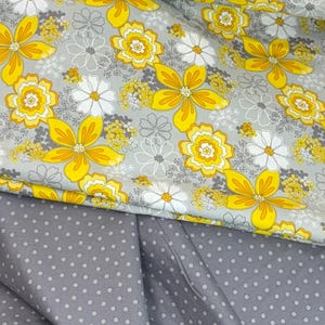 Puede incluir: Primer plano de tela con estampado floral. La capa superior presenta grandes flores amarillas con detalles blancos y grises sobre un fondo gris. La capa inferior es gris con lunares blancos. Ideal para costura.