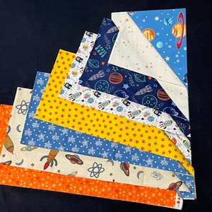 May include: A stack of six fabric squares with a space theme. The squares are in various colors, including blue, yellow, orange, and white. The fabric features stars, planets, rockets, and the words "Space Race".
