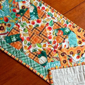 May include: A quilted table runner featuring a patchwork design with autumn-themed patterns. The runner showcases sunflowers, pumpkins, and fall leaves in shades of orange, green, and teal. The edges are trimmed with a striped border.