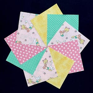 May include: A set of nine fabric squares featuring a pink and white floral pattern, a green polka dot pattern, and a yellow pattern with a subtle shimmer. Each square features a small illustration of two children holding hands with a white goose.