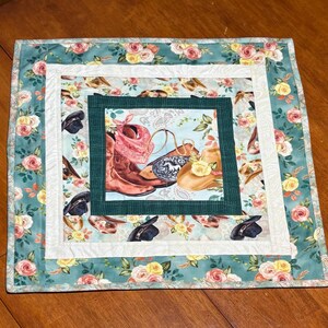 May include: A square quilted placemat with a floral and cowboy theme. The center features a light blue square with cowboy boots, a hat, and a bandana. The border has a floral pattern with roses and a teal background.