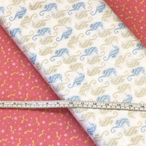 Op de afbeelding: Twee stofstalen met een zeepaardjespatroon. De bovenste staal is wit met blauwe en beige zeepaardjes. De onderste staal is roze met gouden zeepaardjes en witte sterren. Een meetlint ligt op de stalen.