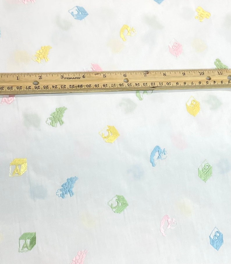 May include: White fabric with a repeating pattern of colourful embroidered blocks and animals. The blocks are in shades of green, yellow, blue, and pink. The animals are a light blue fish, a green dinosaur, and a pink bunny.