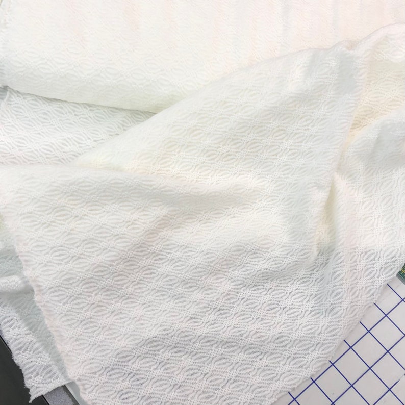May include: White fabric with a textured, woven pattern. The fabric has a subtle sheen and a soft, drapey feel.