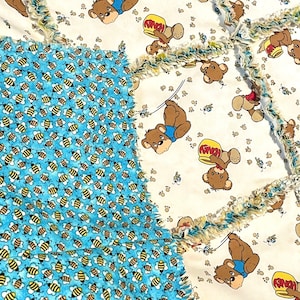 May include: A patchwork quilt featuring a repeating pattern of brown teddy bears with honey pots and bees on a white and light blue background. The quilt has a frayed edge.