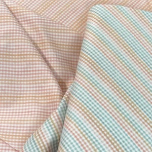 May include: Close-up of folded fabric in pastel colors. The fabric features a gingham pattern in peach and white, and a striped pattern in blue, tan, and white. The fabric is likely cotton or a similar material.