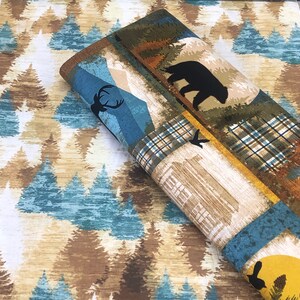 May include: Rolled fabric with a nature-themed design. The fabric features a repeating pattern of evergreen trees in shades of brown, blue, and beige. The rolled section displays panels with a bear, deer, and cabin illustrations, along with plaid and mountain scenes.