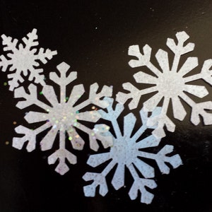 May include: Five white snowflake-shaped paper cutouts on a black background. The snowflakes are different sizes and have a glittery finish.
