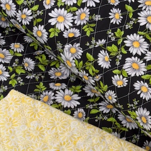 May include: Close-up of two fabrics. One is black with a white daisy pattern, green leaves, and yellow centers, quilted with white thread. The other is yellow with a white daisy pattern.