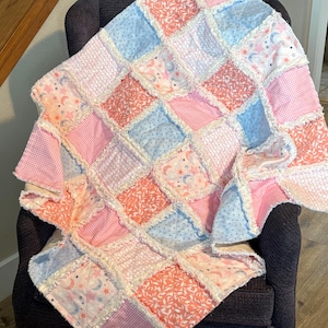 Könnte beinhalten: Eine handgefertigte Patchwork-Decke in Rosa, Hellblau und Koralle, drapiert über einem dunkelgrauen Sessel. Die Decke hat verschiedene gemusterte Quadrate, darunter Vichy-, Blumen- und Himmelsmotive, mit einem Fransenrand.