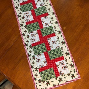 May include: A rectangular Christmas table runner featuring a repeating pattern of red gift boxes with green and white accents. The runner is bordered with a red and white striped trim and holly and berry motifs.
