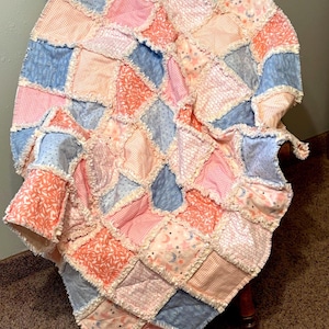 Peut inclure: Une courtepointe patchwork faite à la main, dans des tons de rose, de bleu et de pêche, drapée sur une chaise en bois. La courtepointe présente une variété de tissus à motifs, notamment des motifs floraux, à pois et célestes. Les bords ont une bordure effilochée.
