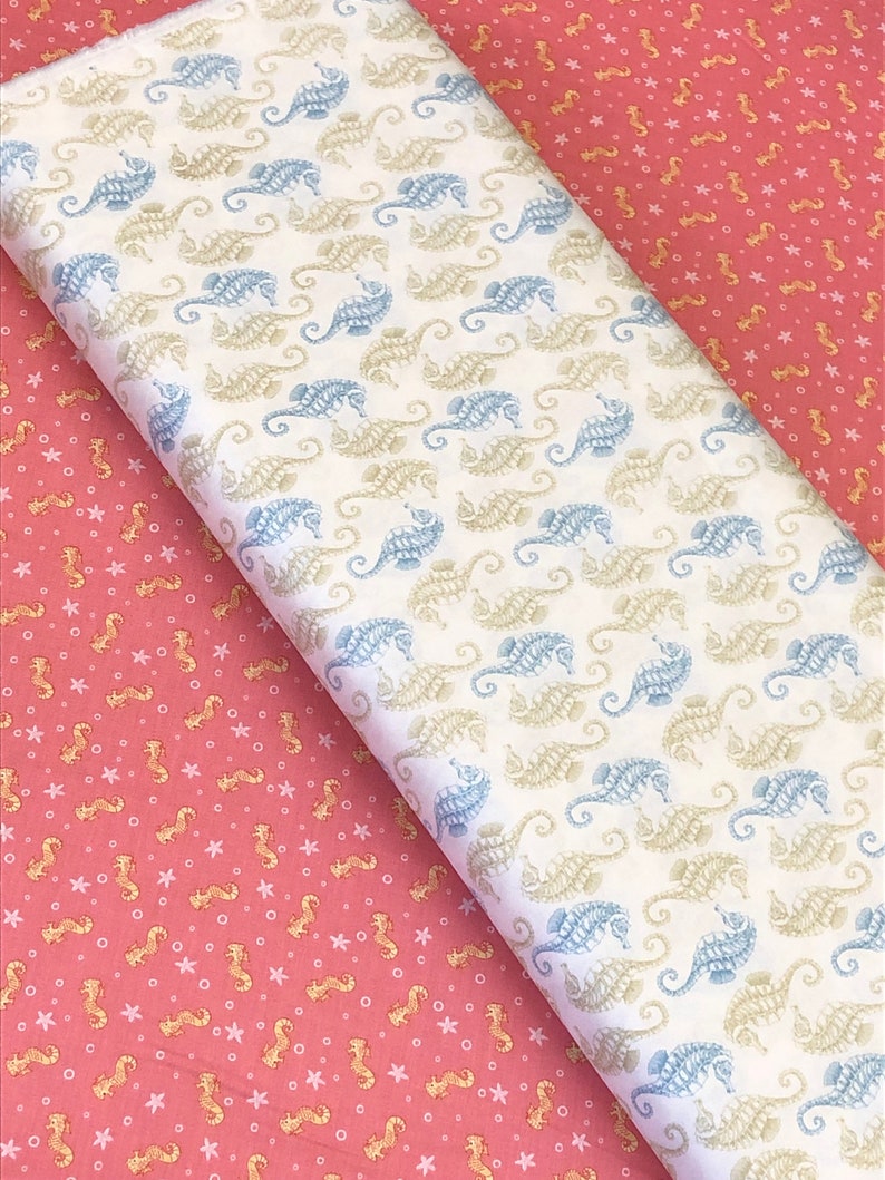 Op de afbeelding: Twee stofstalen met een zeepaardjespatroon. Een staal is wit met blauwe en beige zeepaardjes. De andere staal is roze met gouden zeepaardjes.
