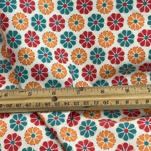 May include: White fabric with a repeating pattern of colorful flowers. The flowers are in shades of red, teal, and orange, with a dark center. A wooden ruler is placed across the fabric, showing inch measurements.