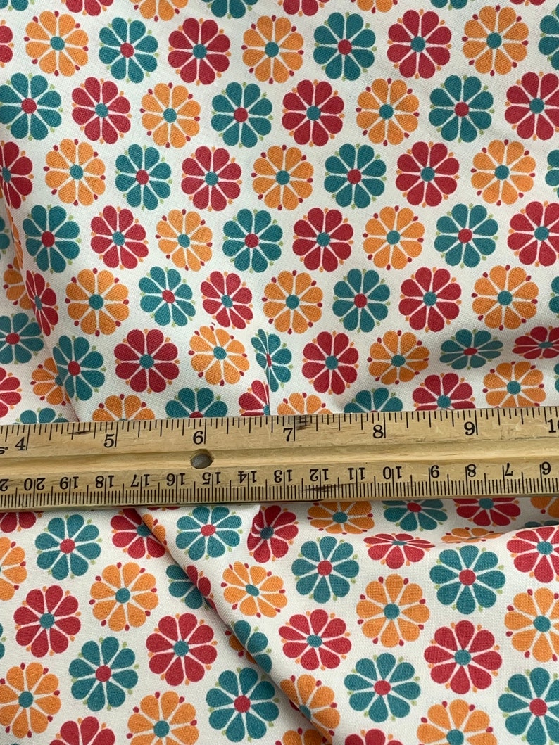 May include: White fabric with a repeating pattern of colorful flowers. The flowers are in shades of red, teal, and orange, with a dark center. A wooden ruler is placed across the fabric, showing inch measurements.