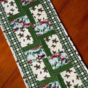 May include: A rectangular Christmas table runner with a patchwork design. The runner features alternating patterns of red trucks in a snowy landscape, holly sprigs, and green plaid. The edges are trimmed with a green plaid pattern.