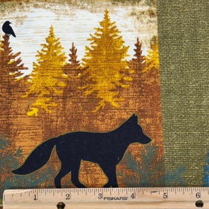 May include: Fabric with a forest scene featuring a black fox silhouette, yellow pine trees, and a black bird. The background has brown, green, and blue tones. A ruler is at the bottom, marked in inches.