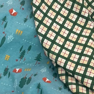 May include: A blue fabric with a repeating pattern of foxes, trees, and mushrooms. A green and white plaid fabric is folded over the blue fabric.
