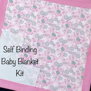 Op de afbeelding: Een roze babydekenset met een zelfbindend ontwerp. De deken heeft een herhalend patroon van roze olifanten en grijze bloemen op een witte achtergrond. De rand is effen roze met een wit stippenpatroon. De tekst "Self Binding Baby Blanket Kit" is zichtbaar.