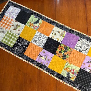 Può includere: Una tovaglia patchwork di Halloween con quadrati di tessuto arancione, nero, viola e giallo. I quadrati mostrano motivi come zucche, pipistrelli e figure di streghe. Il bordo è nero con ragnatele bianche e un bordo a righe.
