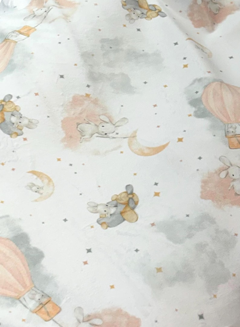 May include: White fabric with a whimsical pattern featuring bunnies, hot air balloons, moons, and stars. The design incorporates soft pink, gray, and gold accents, creating a dreamy, celestial theme. Ideal for nursery decor or baby items.