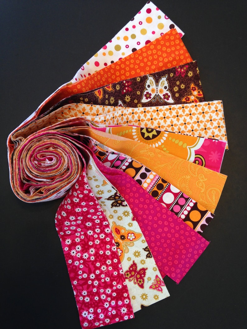 May include: A collection of fabric strips in various shades of orange, pink, and brown with white polka dots, butterfly patterns, and floral designs. The strips are rolled up and fanned out, showcasing the different patterns.