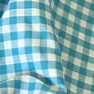 May include: A close-up of a blue and white gingham fabric. The fabric has a checkered pattern with a light blue background and white squares.