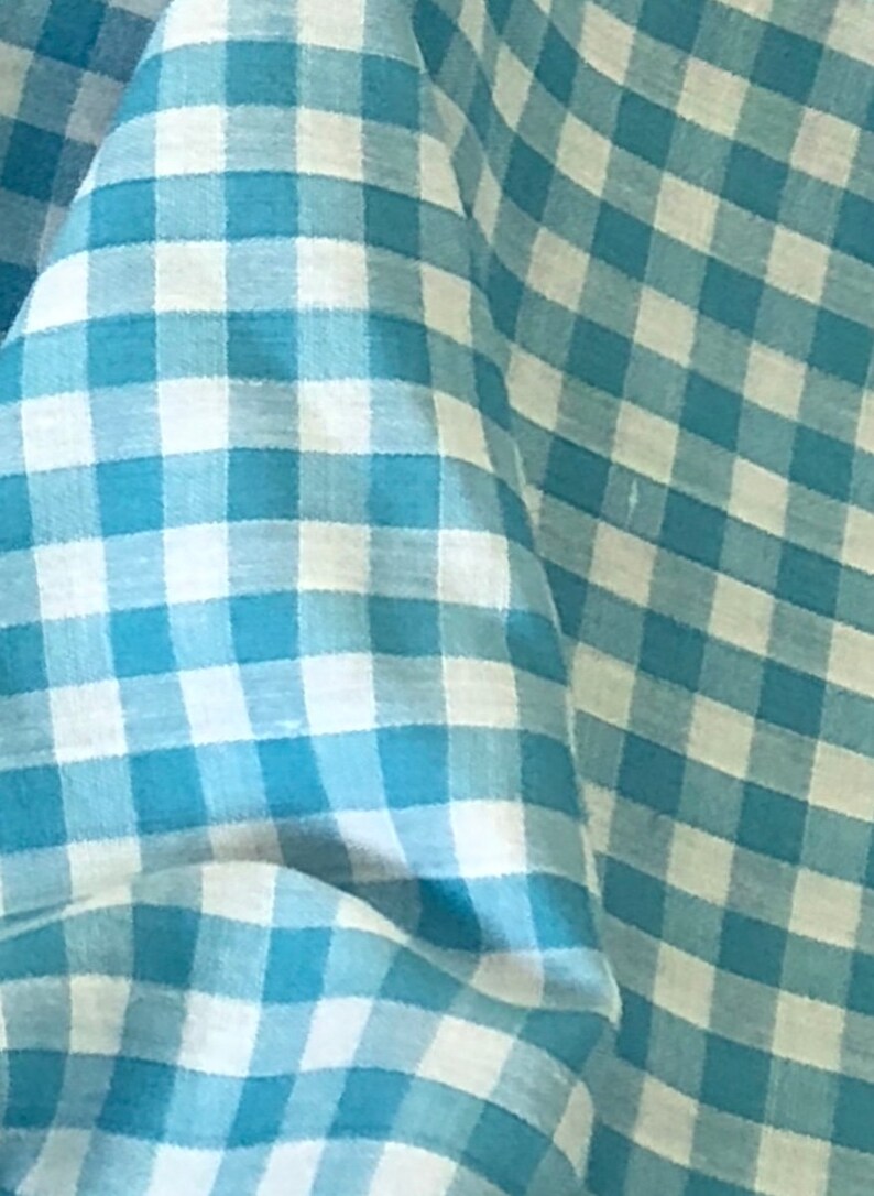 May include: A close-up of a blue and white gingham fabric. The fabric has a checkered pattern with a light blue background and white squares.