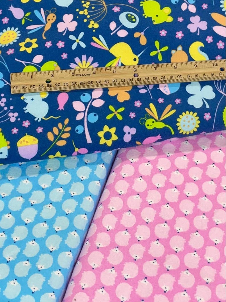 May include: Two fabric swatches with different patterns. The top swatch is blue with a variety of colorful animals, flowers, and insects. The bottom swatch is pink with a repeating pattern of white hedgehogs.