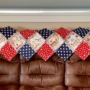 May include: A red, white, and blue patchwork quilt with a frayed edge. The quilt is made up of squares with different patriotic designs, including stars, stripes, and the words "America" and "Freedom".