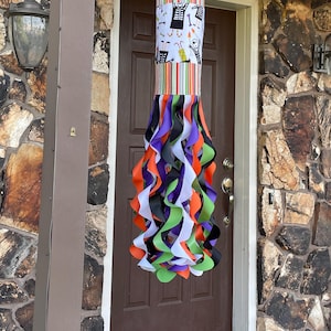 Puede incluir: Una decoración de veleta de Halloween colgada frente a una puerta marrón. La sección superior presenta un panel blanco con figuras de Halloween en blanco y negro y paneles a rayas de colores. La parte inferior tiene largas cintas que fluyen en naranja, morado, negro, verde y blanco.