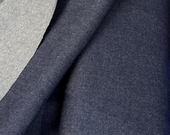 May include: Close-up of a folded piece of dark blue denim fabric. The fabric has a textured surface and is layered over a gray fabric. The denim is a solid color, and the gray fabric is visible in the folds.