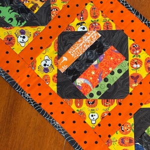 Peut inclure: Chemin de table matelassé sur le thème d'Halloween avec un motif patchwork. Le chemin de table présente des tissus orange, noirs, jaunes et verts avec des motifs d'Halloween comme des crânes, des citrouilles et des araignées. Les bords sont bordés d'un motif de toile d'araignée noir et blanc.