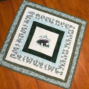 May include: A square quilted table topper with a forest theme. The center features a black bear in front of mountains. The design includes shades of green, cream, and white, with a dotted border. The topper is approximately 24 inches square.